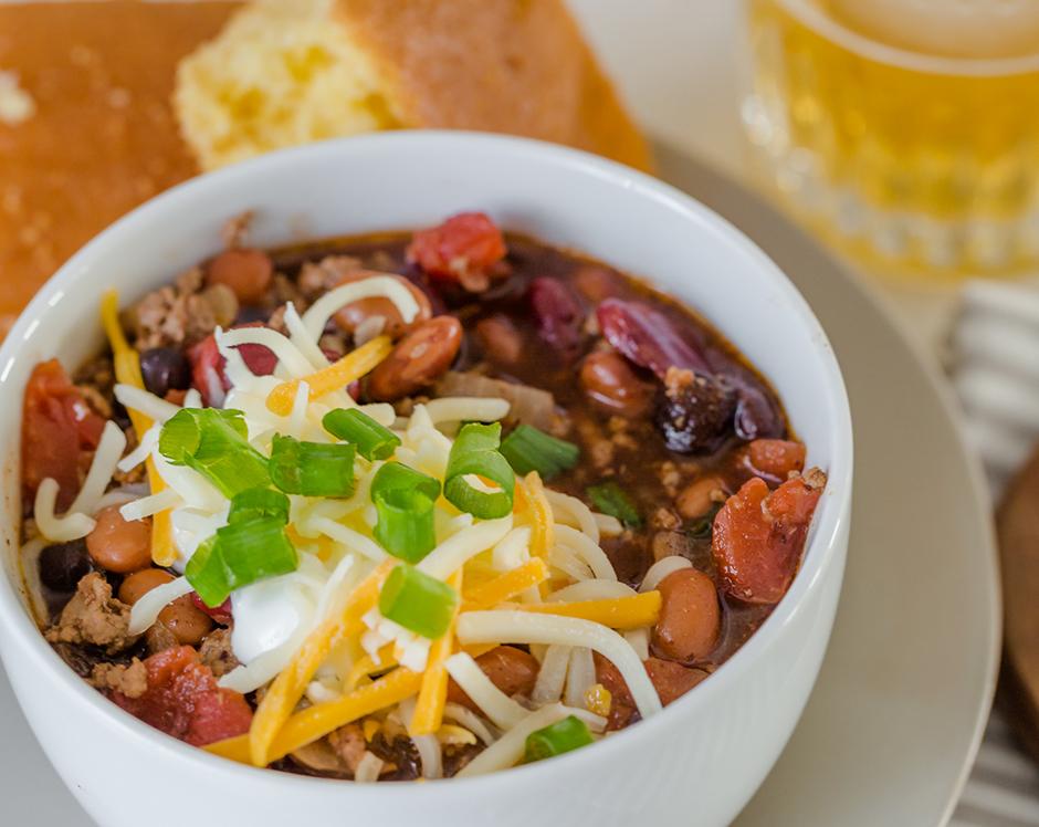 Bowl of chili made with Raven's Nest chili grande mix topped with sour cream, shredded cheese and diced green onions, served with cornbread and honey