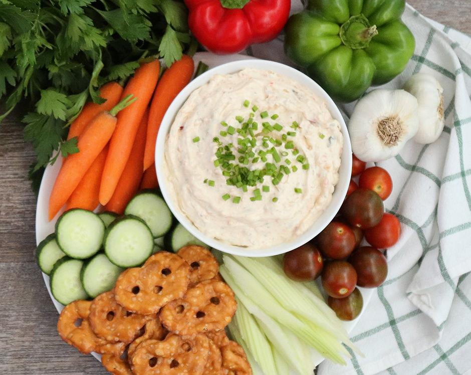 Garden party vegetable dip surrounded by pretzel thins, cucumbers, carrots, bell peppers, garlic, tomatoes, parsley, and celery. Topped with chives.